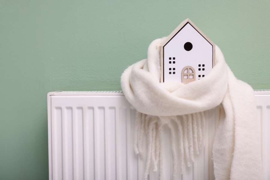 Une maquette de maison enroulée dans une écharpe blanche posée sur un radiateur blanc, symbole d'une maison difficile à chauffer en hiver.