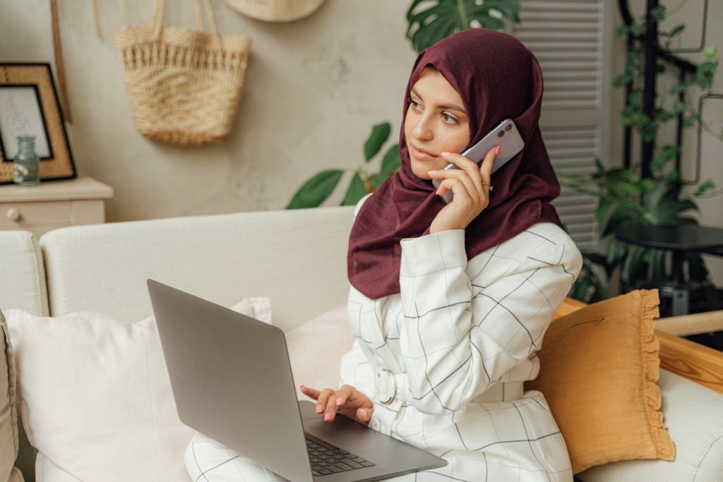 Une jeune femme portant un hijab bordeaux travaille sur son ordinateur portable tout en passant un appel téléphonique, illustrant l'essor de l'IA digital Maroc.