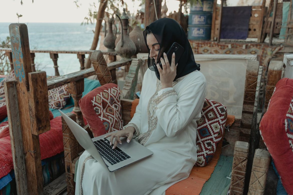 Une femme marocaine en tenue traditionnelle sur une terrasse au bord de l'eau, utilisant un ordinateur portable et son téléphone pour gérer des campagnes de Digital Marketing au Maroc.