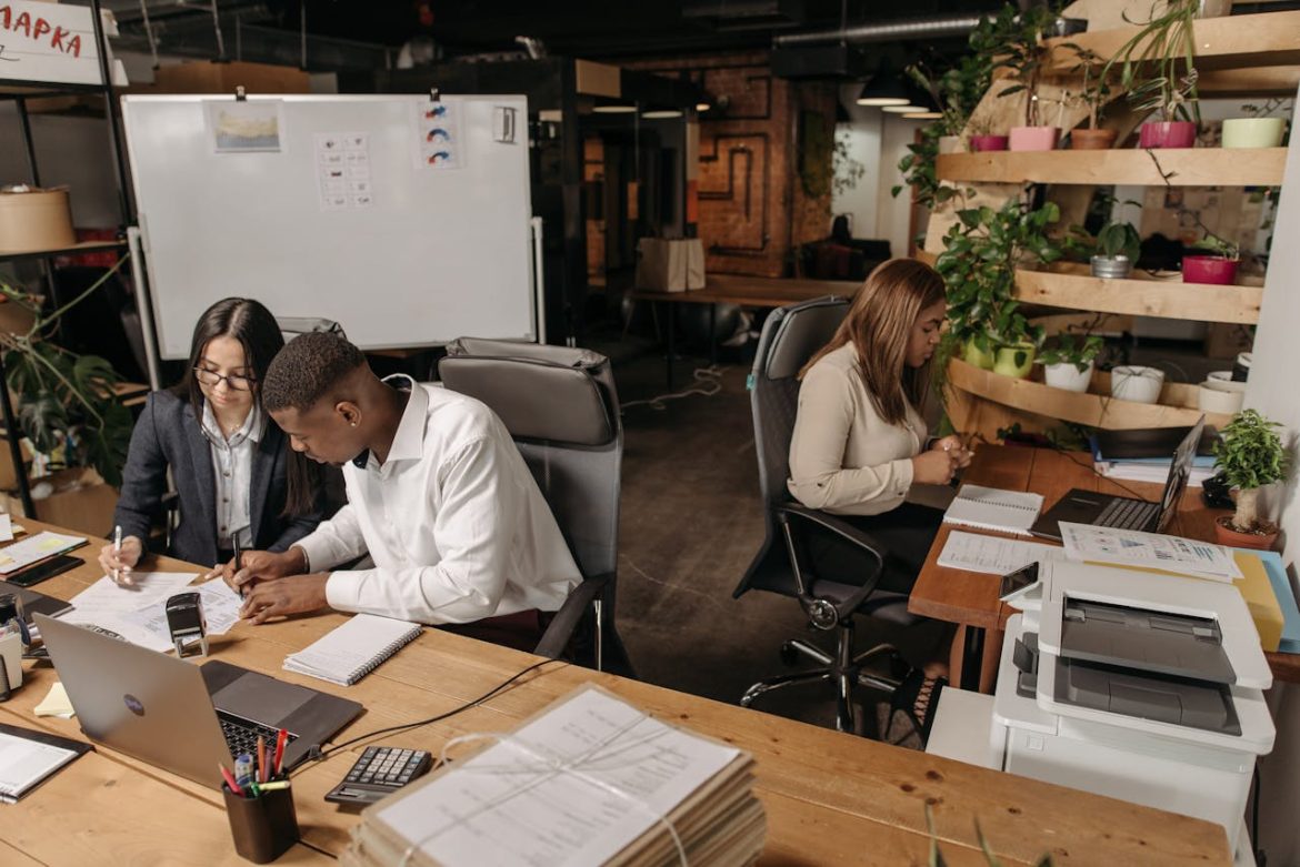 Trois professionnels concentrés travaillant sur des documents et des ordinateurs portables dans un bureau partagé moderne et végétalisé, illustrant comment réussir la transformation digitale de son entreprise au Maroc avec l'IA.