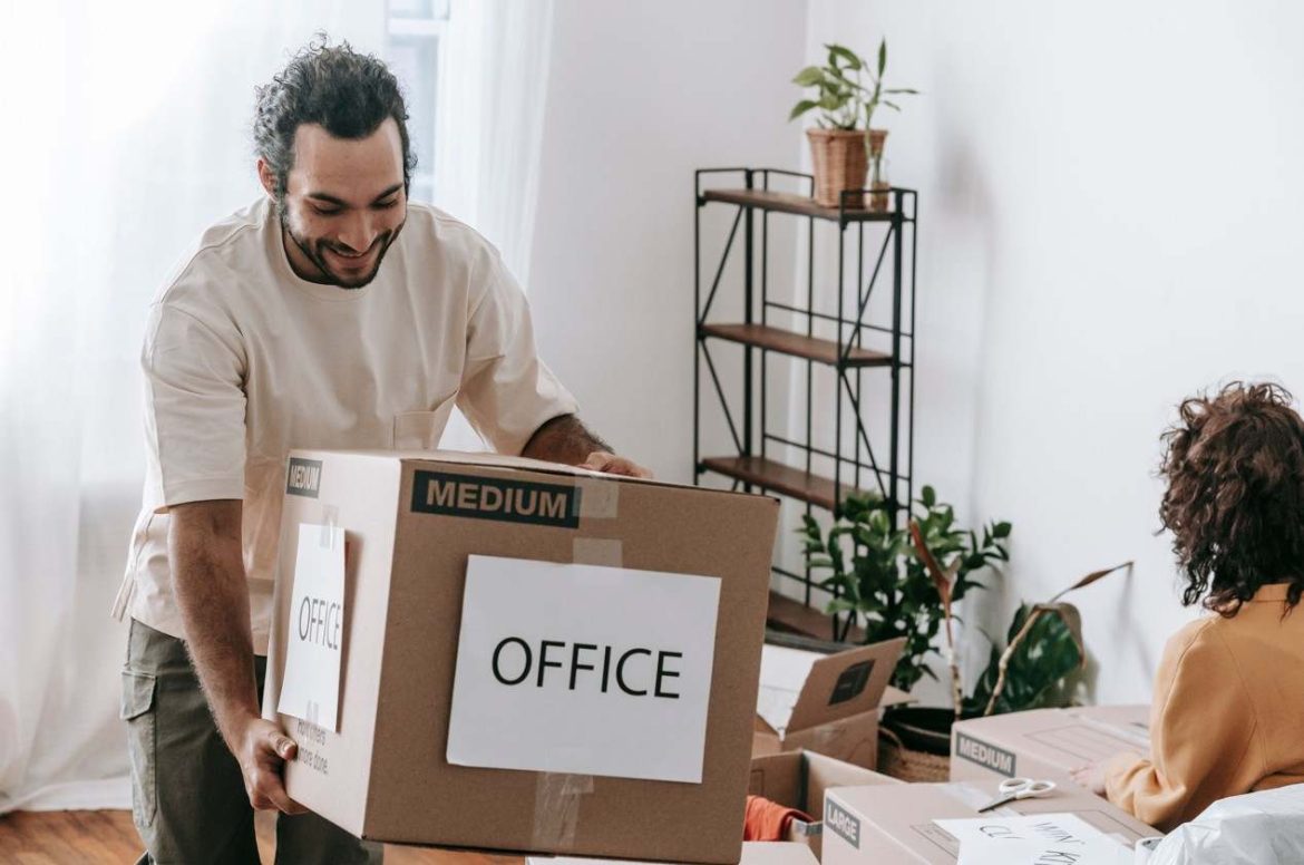 Un collaborateur souriant porte un grand carton étiqueté OFFICE pendant un déménagement de bureau.