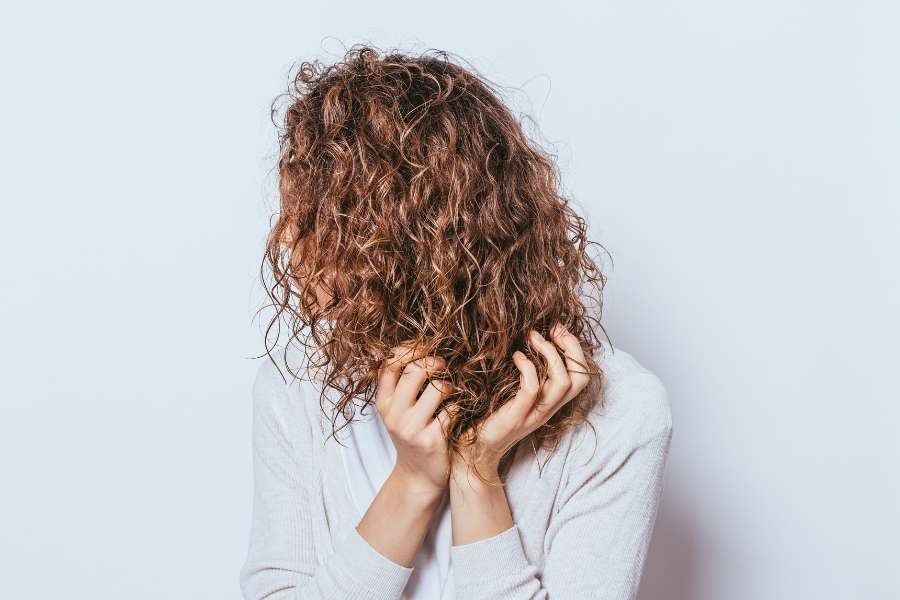 Une femme aux longs cheveux bouclés châtains se tient de profil en touchant ses boucles avec ses mains sur un fond blanc.