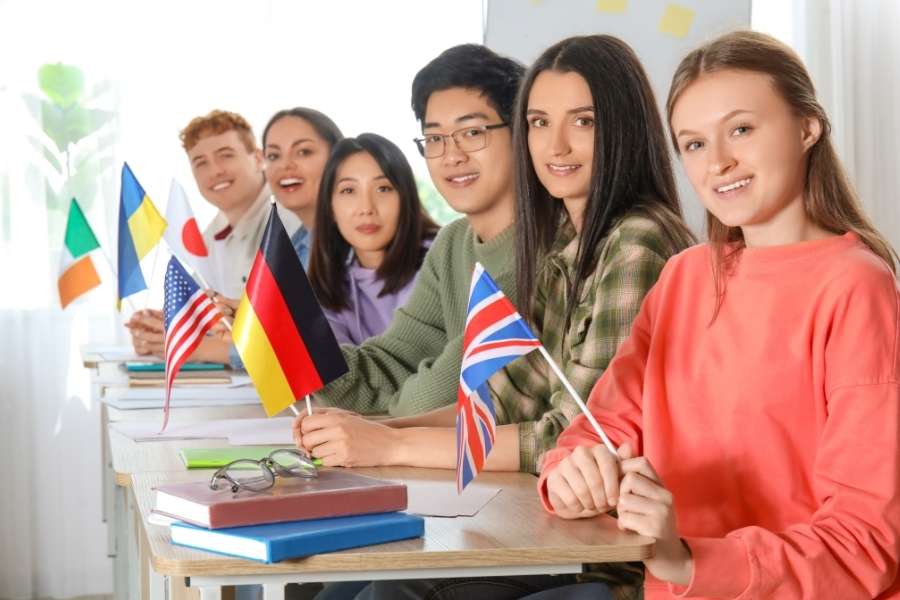 Un groupe d'étudiants internationaux souriants est assis à des bureaux en classe, tenant chacun un petit drapeau pour représenter l'apprentissage d'une langue étrangère.