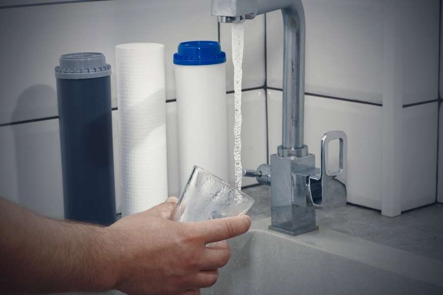 Une main tient un verre sous un robinet d'où coule de l'eau filtrée par un système de traitement de l'eau visible en arrière-plan.